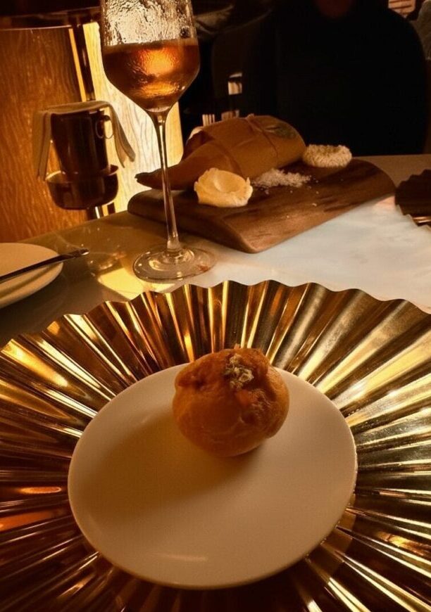 A close-up of a delicate amuse-bouche served on a small white plate, resting atop a large, reflective gold-pleated decorative charger. In the background, a chilled glass of rosé wine and a rustic bread service on a wooden board are illuminated by warm, moody restaurant lighting.