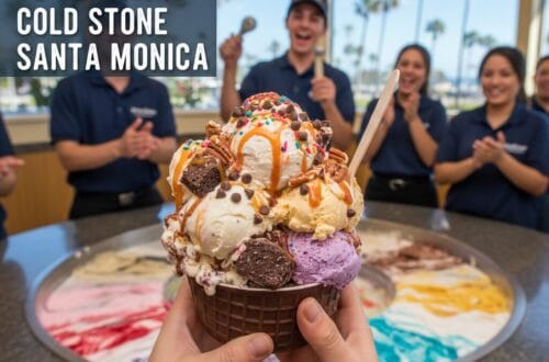 A massive, multi-scoop Cold Stone Creamery ice cream creation in a chocolate waffle bowl with brownies, pecans, and caramel, held over a granite mixing stone while staff cheer in the background.