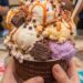 A massive, multi-scoop Cold Stone Creamery ice cream creation in a chocolate waffle bowl with brownies, pecans, and caramel, held over a granite mixing stone while staff cheer in the background.