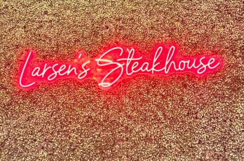 A glowing red neon sign that reads "Larsen’s Steakhouse" mounted on a textured, gold-glittered accent wall at the Woodland Hills location.
