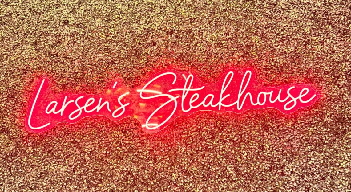 A glowing red neon sign that reads "Larsen’s Steakhouse" mounted on a textured, gold-glittered accent wall at the Woodland Hills location.