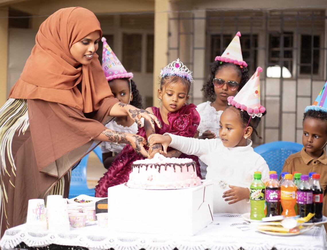 A group of young children in birthday party hats celebrating around a birthday cake