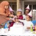 A group of young children in birthday party hats celebrating around a birthday cake