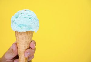 A hand holds a waffle cone topped with a scoop of blue ice cream against a yellow background.