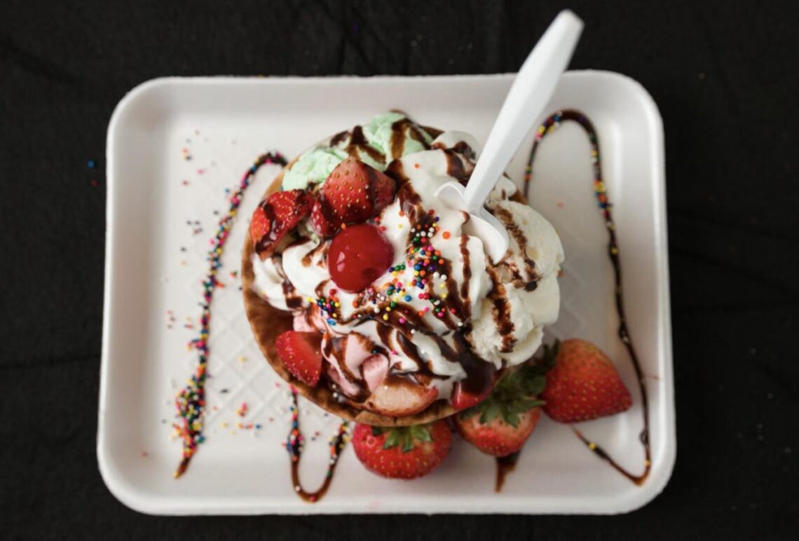 An overhead view of an ice cream sundae topped with whipped cream, chocolate sauce, sprinkles, a cherry, and fresh strawberries.