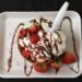 An overhead view of an ice cream sundae topped with whipped cream, chocolate sauce, sprinkles, a cherry, and fresh strawberries.