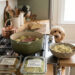 A cozy kitchen counter featuring glass meal prep containers labeled "Sunday: Boil Potatoes" and "Monday: Chop Cabbage" next to a sage green Dutch oven, with a goldendoodle dog peering over the edge.