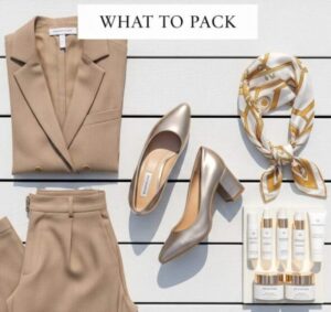 A high-end flat lay of luxury cruise essentials including tan linen trousers, a gold metallic block heel, a silk designer headscarf, and high-end travel skincare on a white wooden deck.