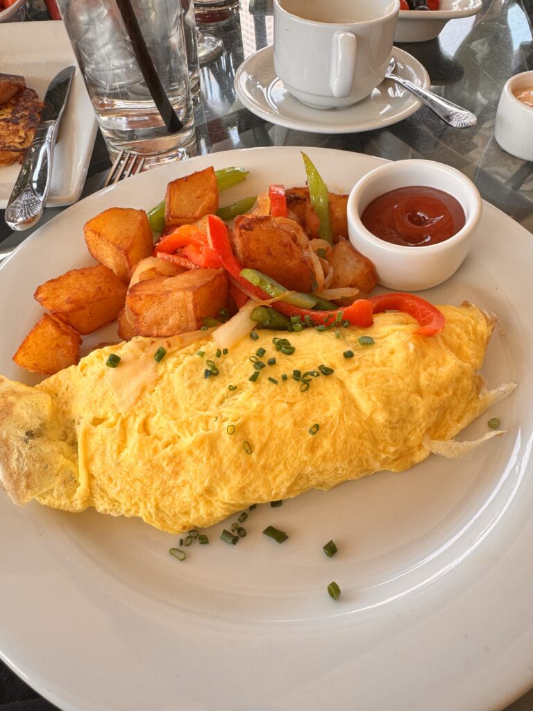A classic French omelet topped with chives, served with crispy breakfast potatoes and sautéed peppers on a white plate at Perch LA.