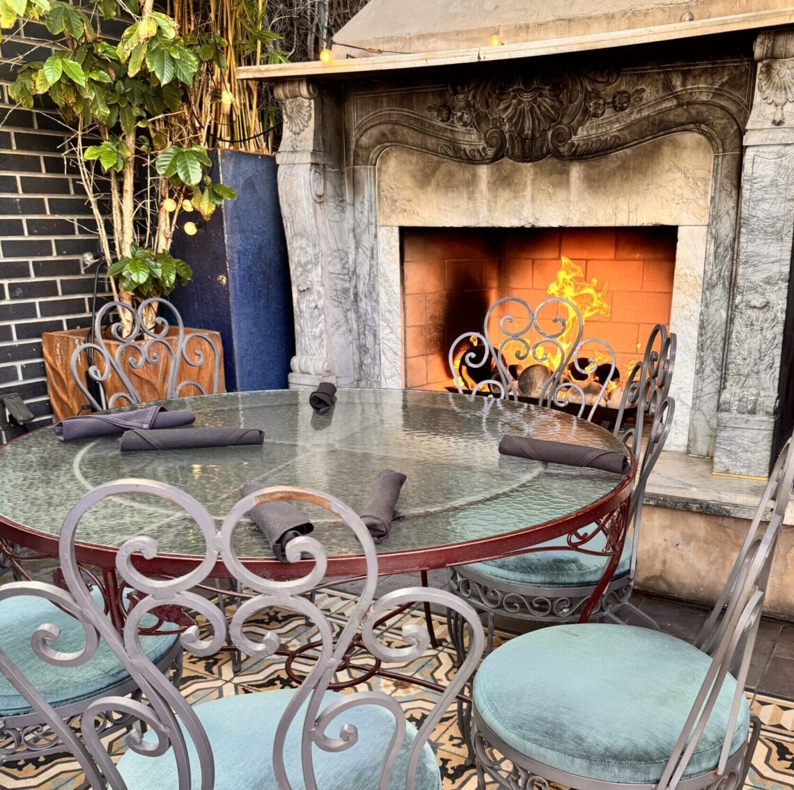 A cozy outdoor patio at Perch LA featuring a roaring stone fireplace and an ornate wrought-iron table and chairs on geometric patterned tile.
