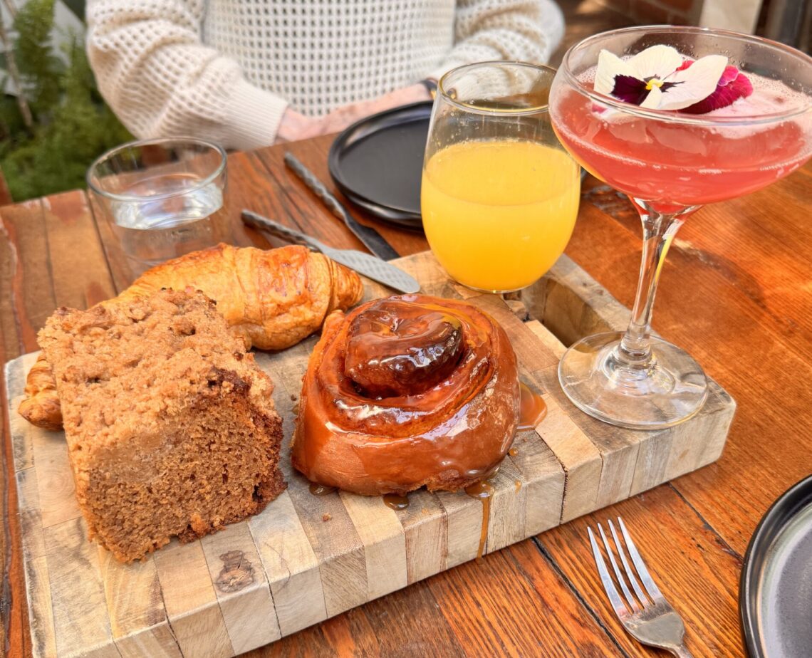 A wooden board featuring a large cinnamon roll, a croissant, and a slice of coffee cake at Fia Santa Monica, accompanied by orange juice and a pink floral cocktail.
