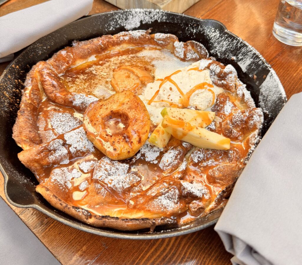 A large, golden-brown Dutch Baby pancake served in a black cast iron skillet at Fia Santa Monica, topped with roasted apples, powdered sugar, and a scoop of melting cream on a rustic wooden table.