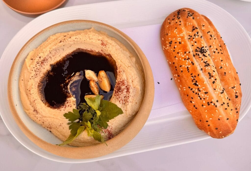 Overhead view of a creamy hummus bowl topped with dark oil, whole roasted garlic cloves, and fresh herbs, served alongside a toasted Persian sesame bread roll.