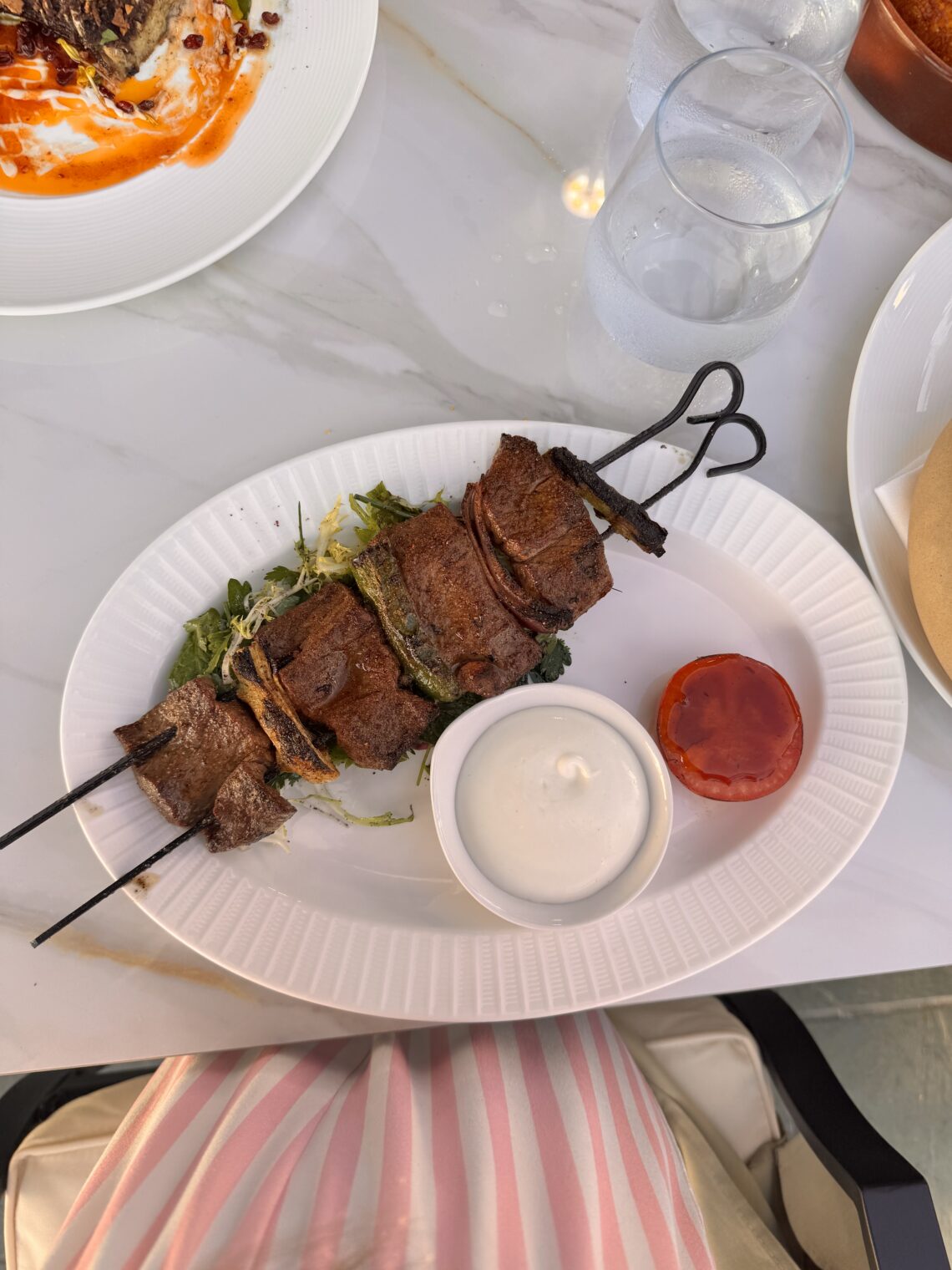 A gourmet plate featuring a Filet Mignon Kabob on a black metal skewer with a side of white creamy sauce and a roasted tomato slice at Perse in Brentwood.