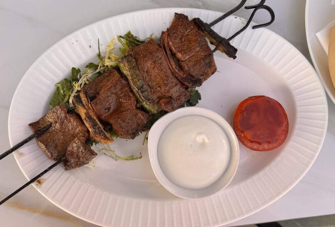 A gourmet plate featuring a Filet Mignon Kabob on a black metal skewer with a side of white creamy sauce and a roasted tomato slice at Perse in Brentwood.