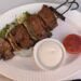 A gourmet plate featuring a Filet Mignon Kabob on a black metal skewer with a side of white creamy sauce and a roasted tomato slice at Perse in Brentwood.