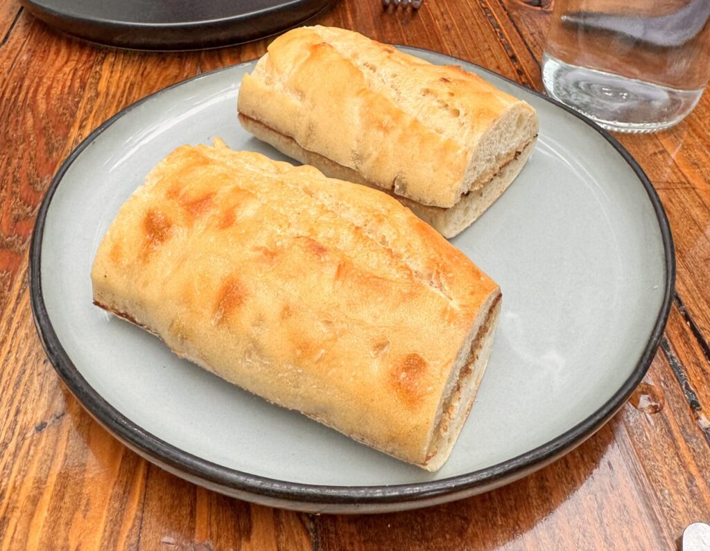 Two thick, golden-brown toasted garlic baguette slices served on a light blue ceramic plate with a dark rim, resting on a rustic wooden dining table at Fia Santa Monica.