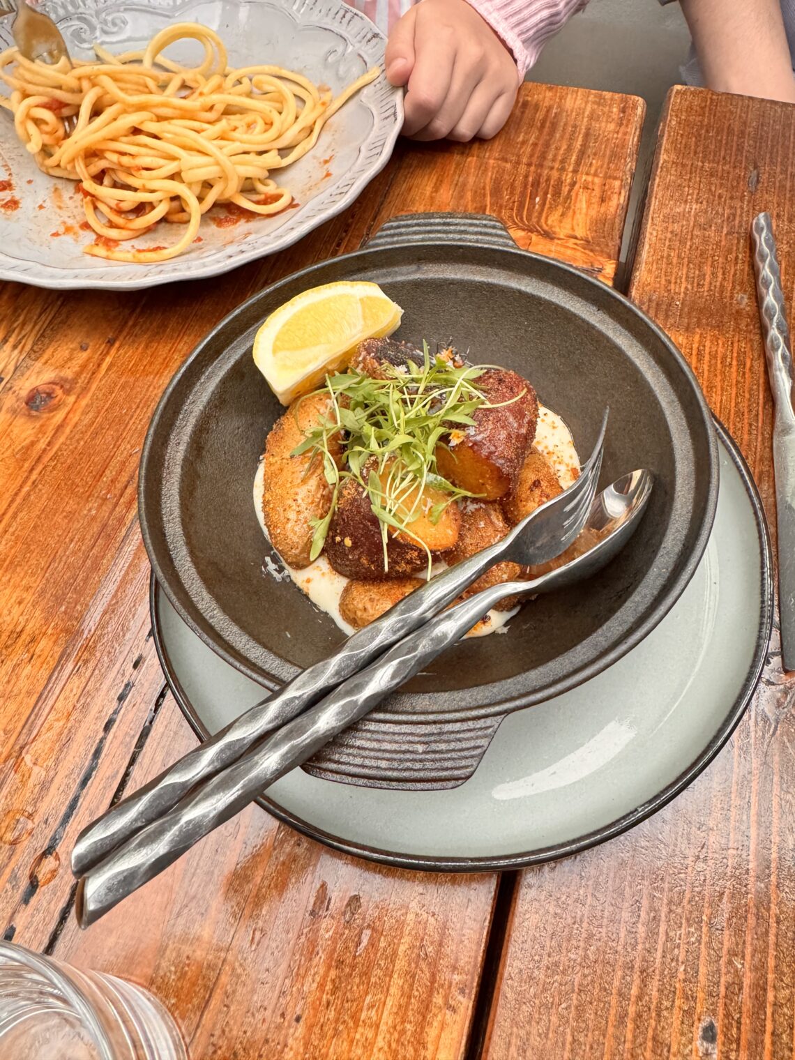 An overhead photograph of golden roasted potatoes in a dark rustic ceramic serving bowl with a handle, garnished with fresh green microgreens, sea salt flakes, and coarse black pepper. A large lemon wedge rests on the rim. The bowl sits on a weathered wooden patio table with hammered metal serving spoon and fork.