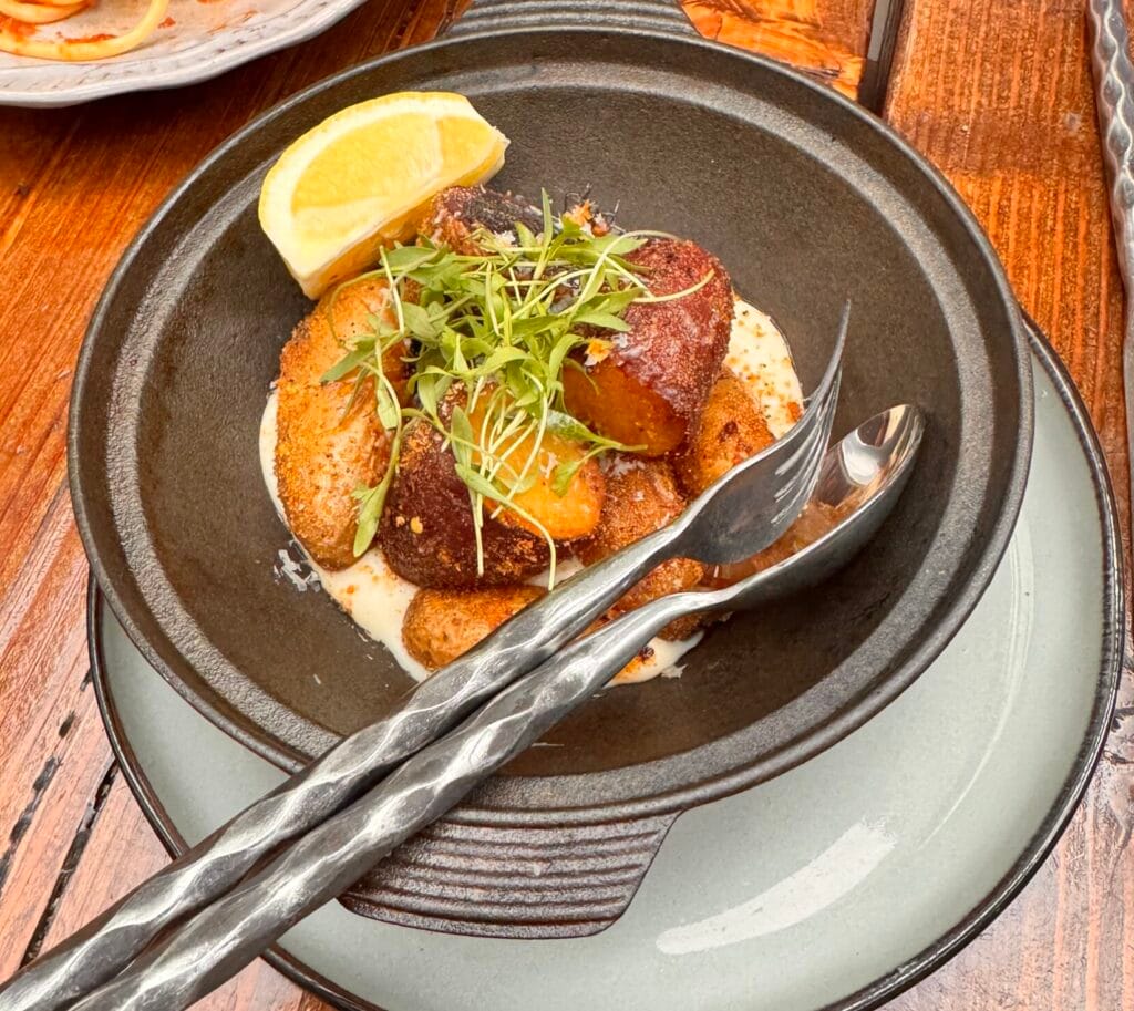 An overhead photograph of golden roasted potatoes in a dark rustic ceramic serving bowl with a handle, garnished with fresh green microgreens, sea salt flakes, and coarse black pepper. A large lemon wedge rests on the rim. The bowl sits on a weathered wooden patio table with hammered metal serving spoon and fork.