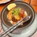 An overhead photograph of golden roasted potatoes in a dark rustic ceramic serving bowl with a handle, garnished with fresh green microgreens, sea salt flakes, and coarse black pepper. A large lemon wedge rests on the rim. The bowl sits on a weathered wooden patio table with hammered metal serving spoon and fork.