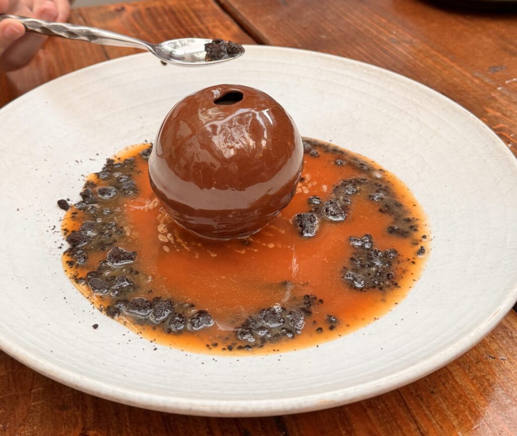 A glossy dark chocolate sphere dessert centered on a white plate with bright orange-citrus sauce and dark chocolate crumbles, being served with a silver spoon at Fia Santa Monica.