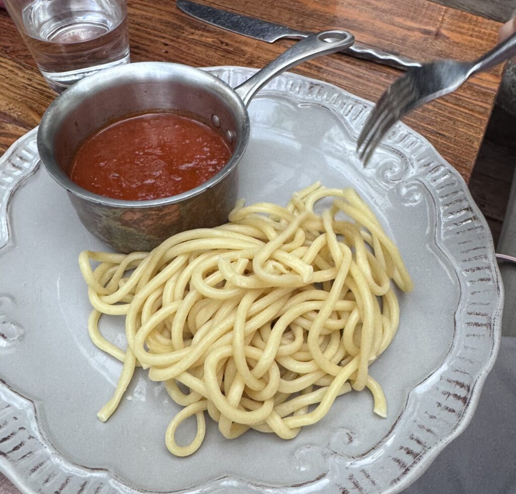 Overhead view of a decorative scalloped white plate holding plain, thick spaghetti-style pasta, served with a miniature stainless steel pot of red marinara sauce on a rustic wooden table, as a hand with a fork prepares to take a bite.
