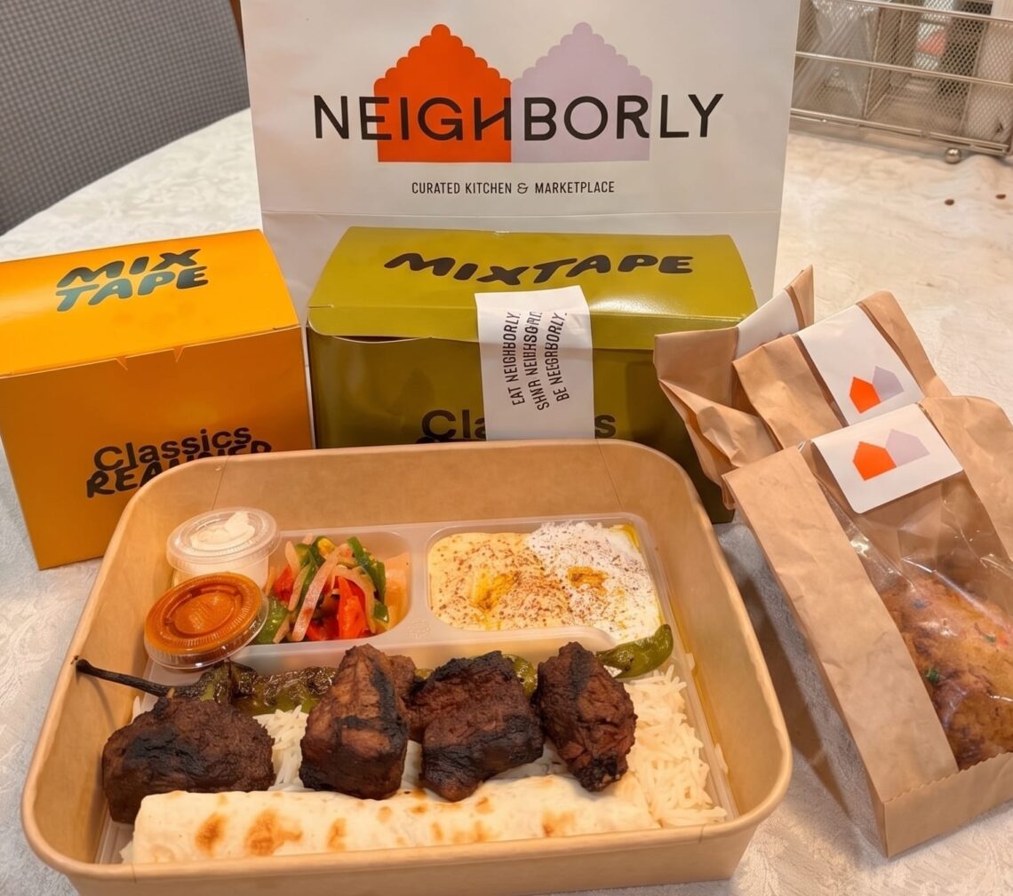 An overhead photograph on a white tablecloth unboxing a family delivery feast from Neighborly Kitchen and Marketplace Brentwood. The segmented container in the foreground holds a large portion of flame-charred Mini Kabob steak, rice, hummus, and salad. Behind it, the yellow "MIX TAPE" box and green "CLASSICS" boxes are clearly visible, next to two brown paper treat bags with small orange and white roof-shaped stickers from Neighborly Kitchen.