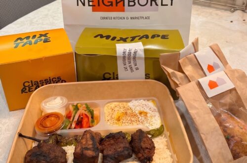 An overhead photograph on a white tablecloth unboxing a family delivery feast from Neighborly Kitchen and Marketplace Brentwood. The segmented container in the foreground holds a large portion of flame-charred Mini Kabob steak, rice, hummus, and salad. Behind it, the yellow "MIX TAPE" box and green "CLASSICS" boxes are clearly visible, next to two brown paper treat bags with small orange and white roof-shaped stickers from Neighborly Kitchen.