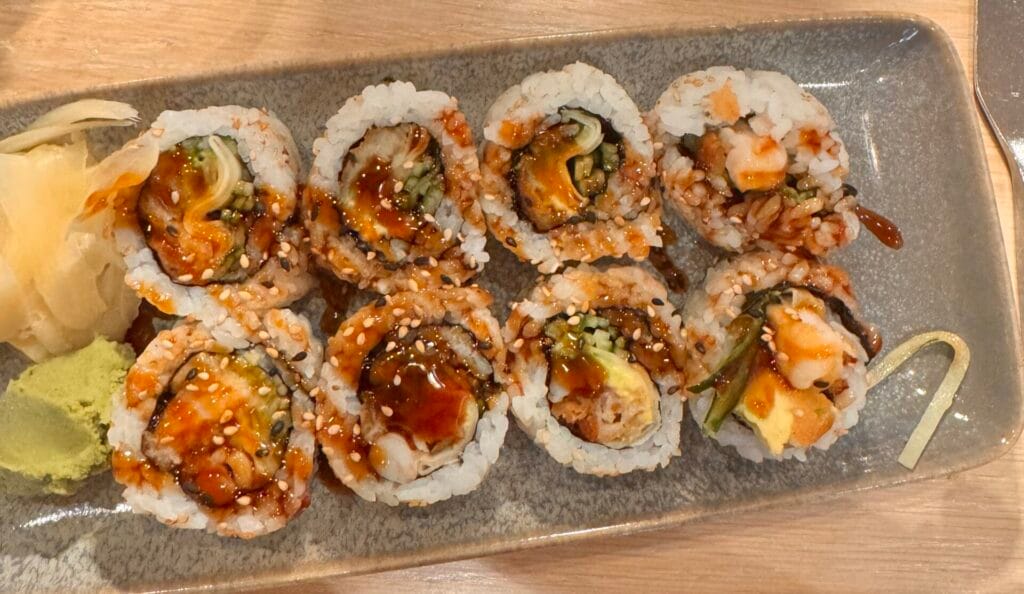 A top-down view of eight pieces of shrimp tempura sushi rolls on a grey rectangular plate with ginger and wasabi at Pacific Catch Santa Monica.