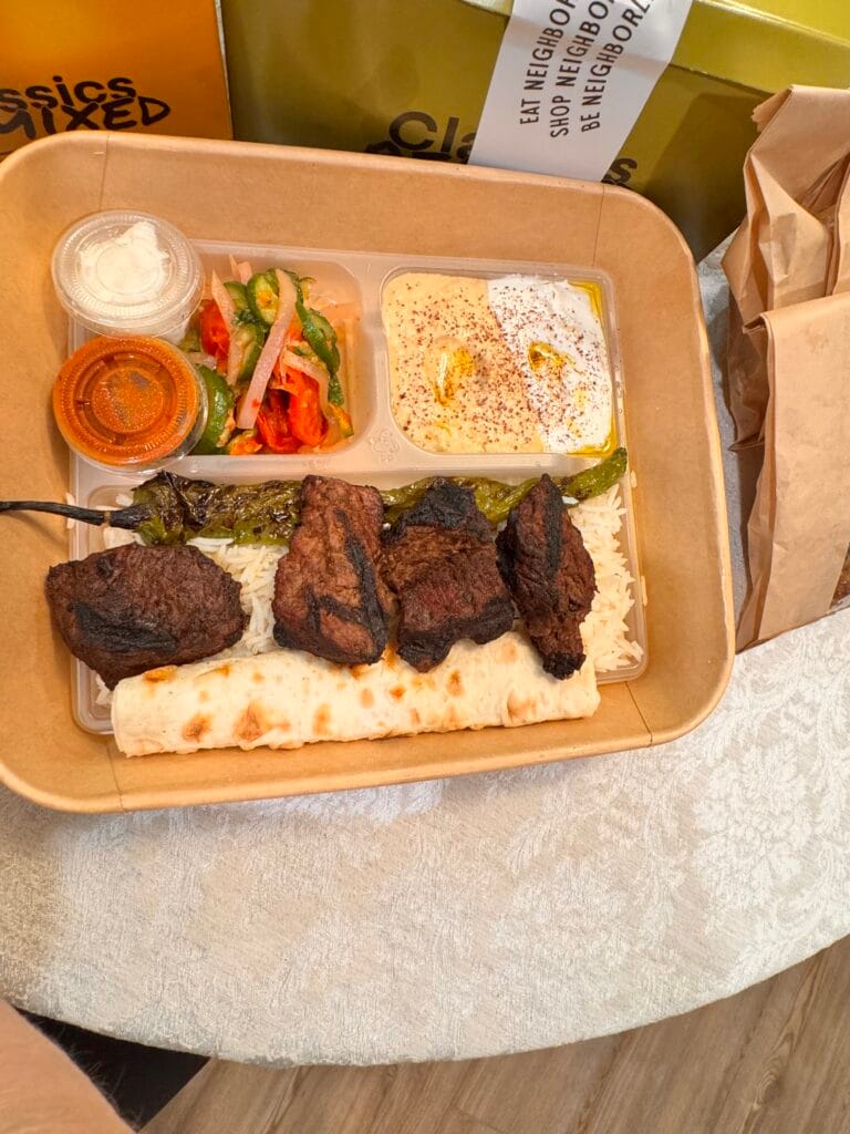 An overhead photograph of the Papa Hov’s Steak Plate from Mini Kabob, delivered in a sustainable craft paper bowl. The meal features four large, flame-broiled marinated steak pieces over basmati rice, a grilled shishito pepper, lavash, hummus with sumac, a vegetable salad, and individual sides of tzatziki mast and spicy red sauce from Neighborly Kitchen Brentwood.
