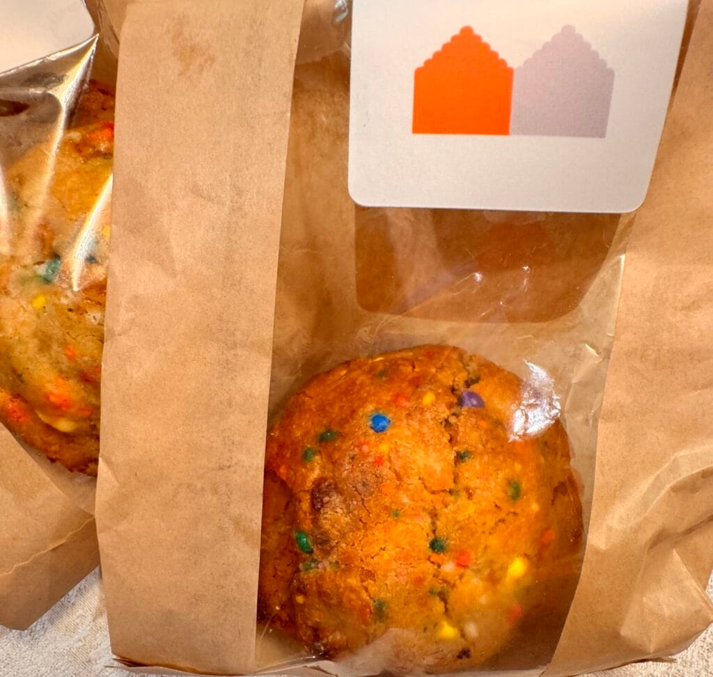 A close-up of a large, golden-brown Birthday Cake cookie with colorful rainbow sprinkles inside a brown paper delivery bag with a transparent window. The bag features the Neighborly Kitchen logo sticker at the top, shown on a white textured tablecloth.