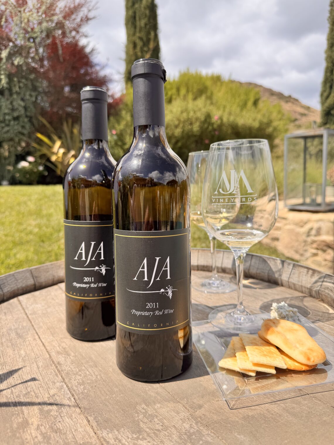 Two bottles of AJA Vineyards 2011 Proprietary Red Wine sitting on a rustic wooden wine barrel alongside etched AJA wine glasses and a small plate of crackers and cheese at the Agoura estate.