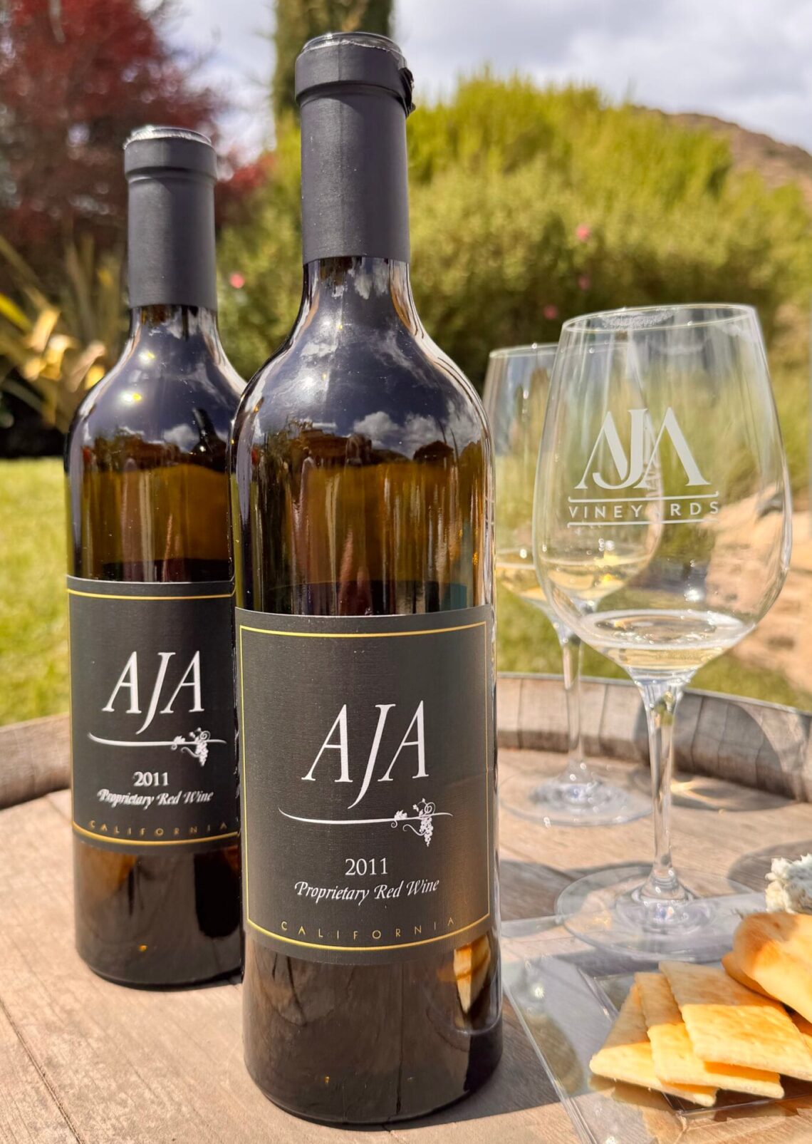 Two bottles of AJA Vineyards 2011 Proprietary Red Wine sitting on a rustic wooden wine barrel alongside etched AJA wine glasses and a small plate of crackers and cheese at the Agoura estate.