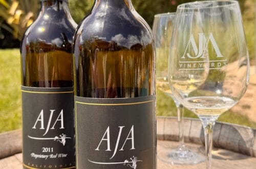 Two bottles of AJA Vineyards 2011 Proprietary Red Wine sitting on a rustic wooden wine barrel alongside etched AJA wine glasses and a small plate of crackers and cheese at the Agoura estate.