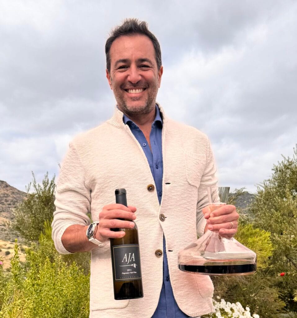 Todd Greenbaum in a cream cardigan and white trousers smiling outdoors at a vineyard, holding a bottle of AJA wine and a glass wine decanter.