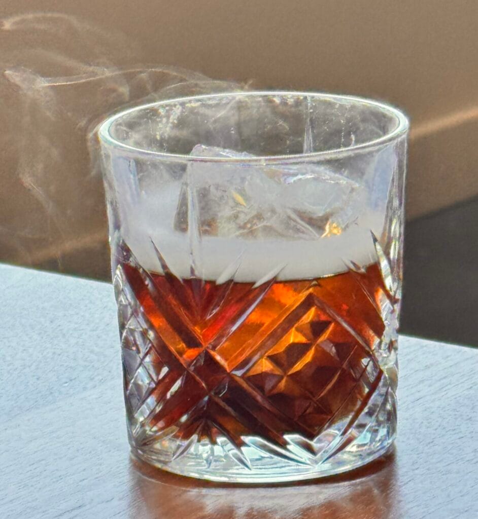 A dark, amber-colored La Sombra Portena cocktail served in a heavy crystal rocks glass with a large square ice cube and a plume of aromatic smoke rising from the surface.