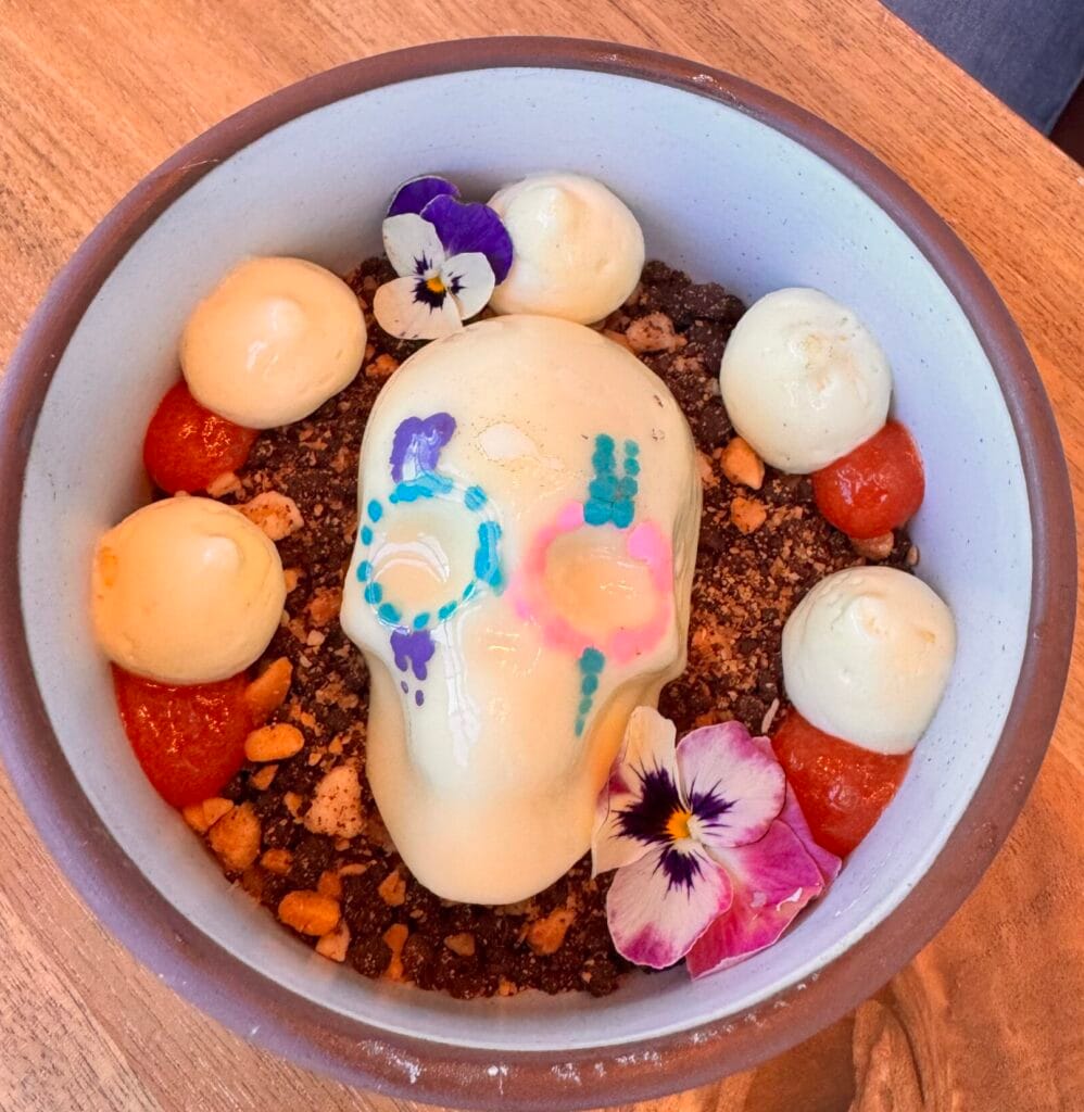 A white chocolate sugar skull dessert in a bowl, decorated with blue and pink edible paint, surrounded by chocolate soil, meringue dollops, red fruit gel, and edible pansy flowers.