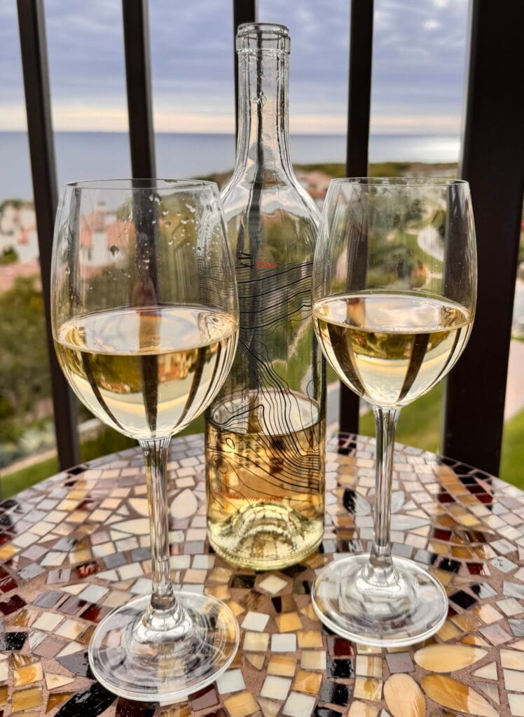 Two glasses of AJA’s 2025 Ed’s Vineyard Sauvignon Blanc Wine and a clear glass bottle with a topographic map design sit on a mosaic table on a balcony overlooking the ocean.
