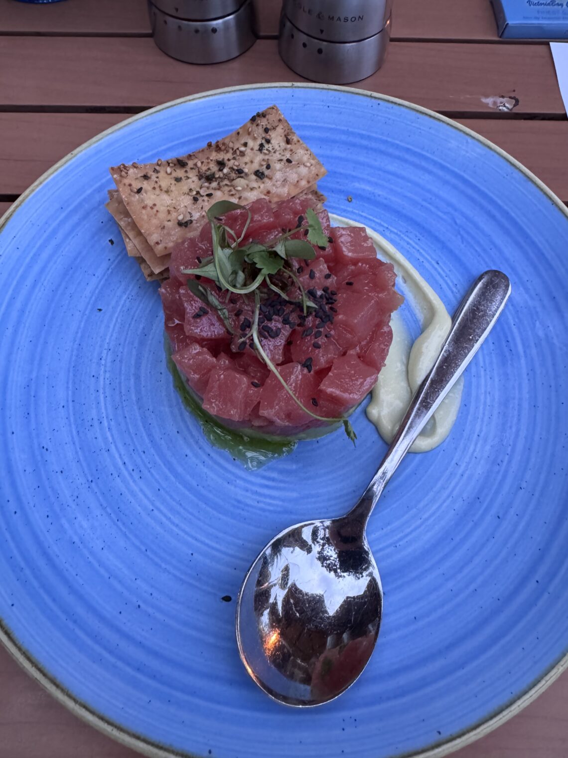 A circular stack of diced raw red tuna topped with sprouts and black sesame seeds, served on a bright blue textured plate with crackers and a side of creamy sauce
