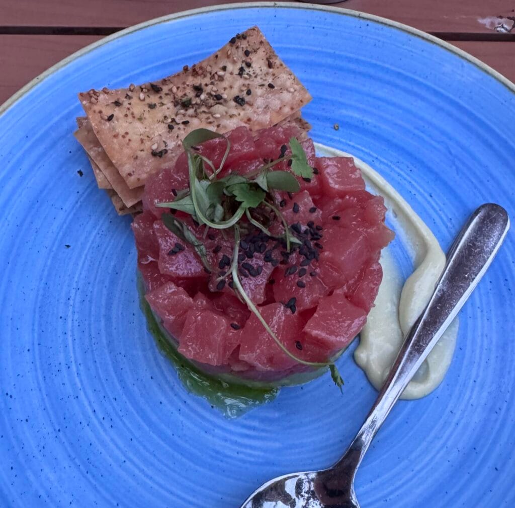 A circular stack of diced raw red tuna topped with sprouts and black sesame seeds, served on a bright blue textured plate with crackers and a side of creamy sauce