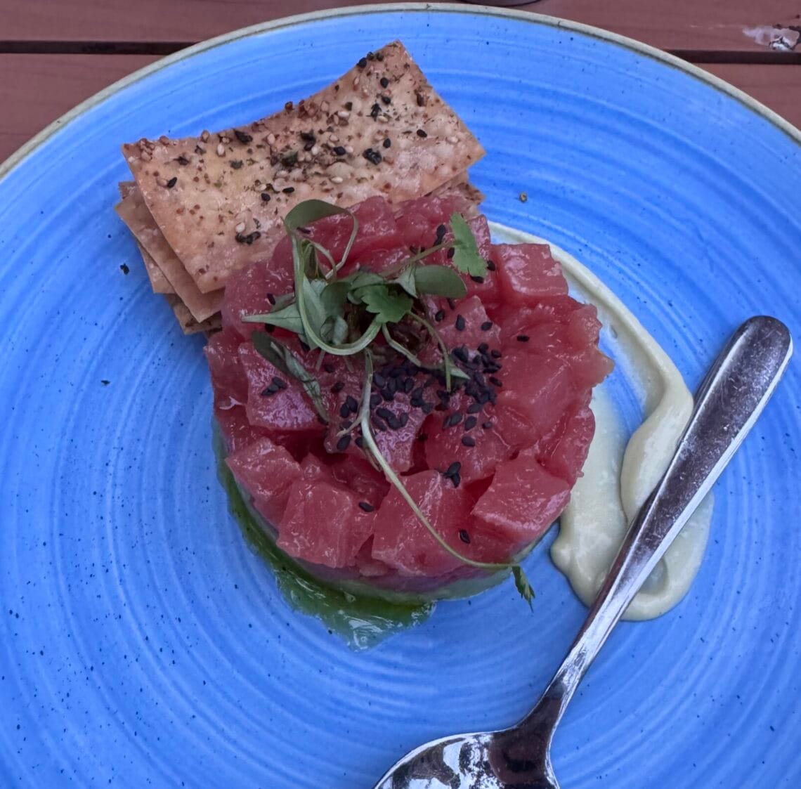 A circular stack of diced raw red tuna topped with sprouts and black sesame seeds, served on a bright blue textured plate with crackers and a side of creamy sauce