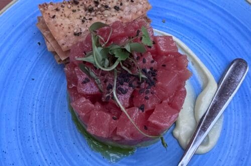 A circular stack of diced raw red tuna topped with sprouts and black sesame seeds, served on a bright blue textured plate with crackers and a side of creamy sauce
