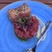 A circular stack of diced raw red tuna topped with sprouts and black sesame seeds, served on a bright blue textured plate with crackers and a side of creamy sauce
