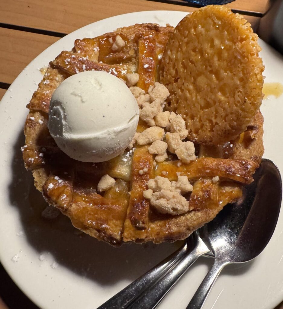 The Terranea Catalina Kitchen small lattice-crust apple pie on a white plate, topped with a scoop of vanilla ice cream, crumble pieces, and a large lace cookie.