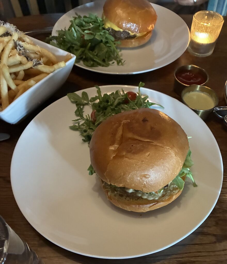 A top-down view of a dinner table setting featuring two gourmet burgers served on white round plates. In the foreground, a large burger with a glossy, golden-brown brioche bun sits on a plate accompanied by a side salad of fresh arugula and cherry tomatoes. In the background, another similar burger is visible next to a glowing candle in a ribbed glass holder. To the left, a white square bowl overflows with golden french fries topped with shaved truffle and parmesan cheese, while two small metal ramekins containing ketchup and a pale yellow dipping sauce sit nearby on the dark wooden table.