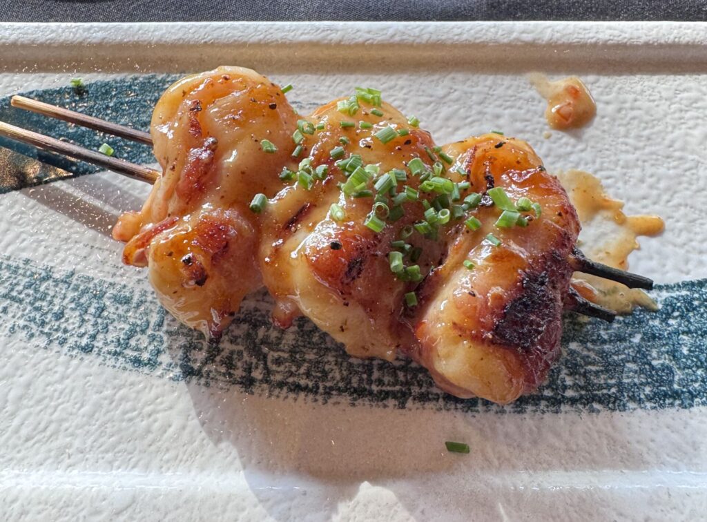Close-up of three average-sized bacon-wrapped shrimp on wooden skewers, drizzled with glaze and topped with chopped chives, served on a textured white and teal ceramic plate at Sushi Roku in Santa Monica.