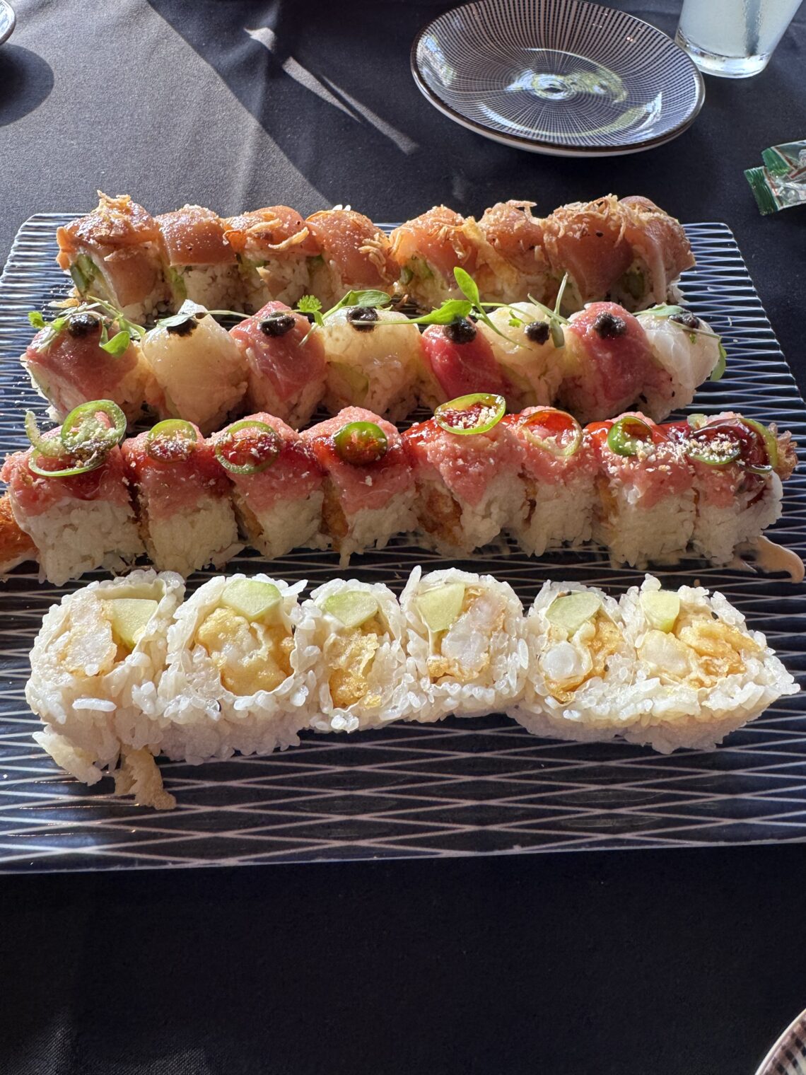 An overhead view of a large platter featuring four varieties of specialty sushi rolls, including the Tokubetsu Roll topped with delicate microgreens and black accents, the White Lotus Roll, a spicy tuna roll with jalapeño slices, and a popcorn shrimp tempura roll, served on a textured blue and white glass plate at Sushi Roku in Santa Monica.
