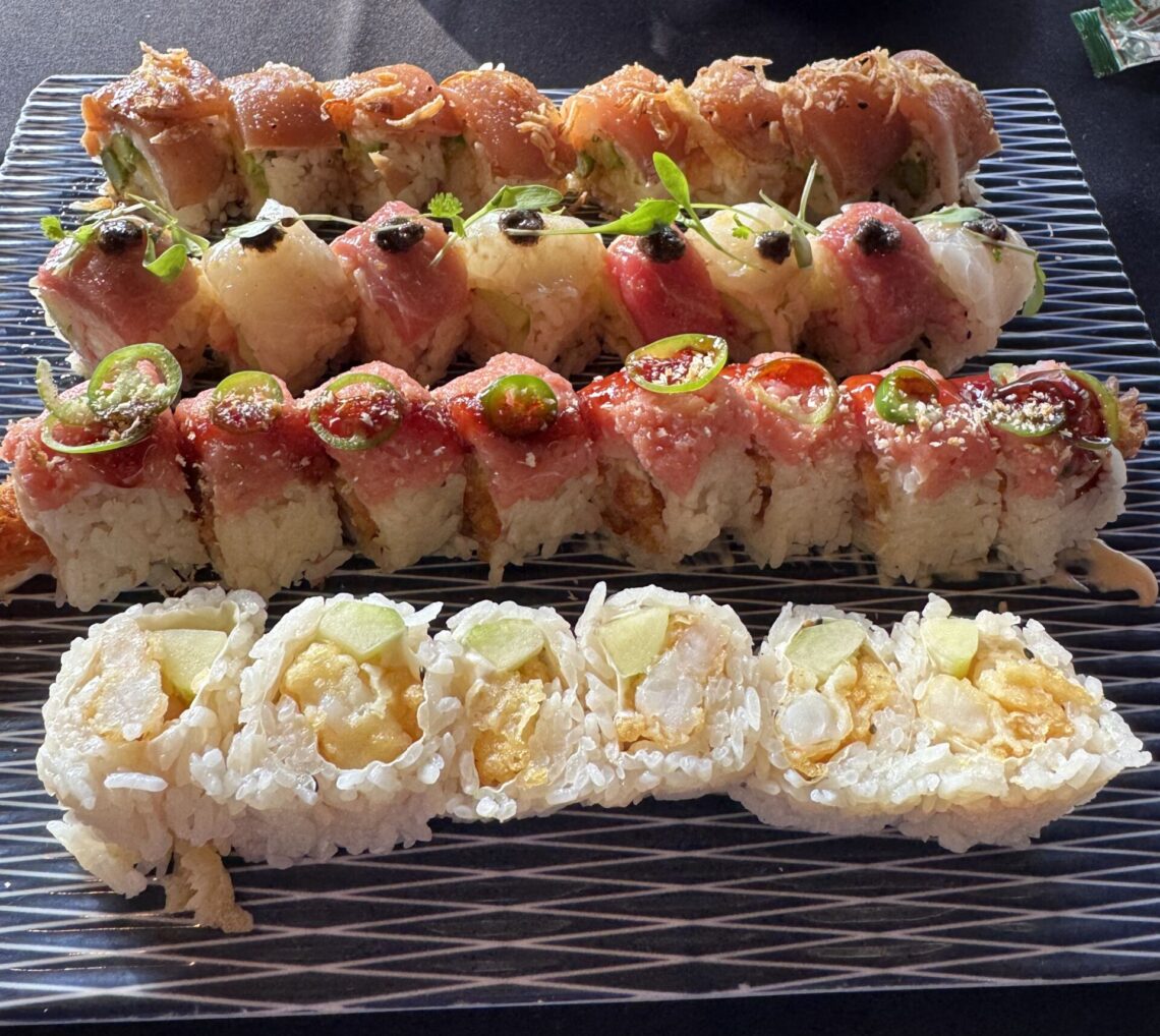 An overhead view of a large platter featuring four varieties of specialty sushi rolls, including the Tokubetsu Roll topped with delicate microgreens and black accents, the White Lotus Roll, a spicy tuna roll with jalapeño slices, and a popcorn shrimp tempura roll, served on a textured blue and white glass plate at Sushi Roku in Santa Monica.