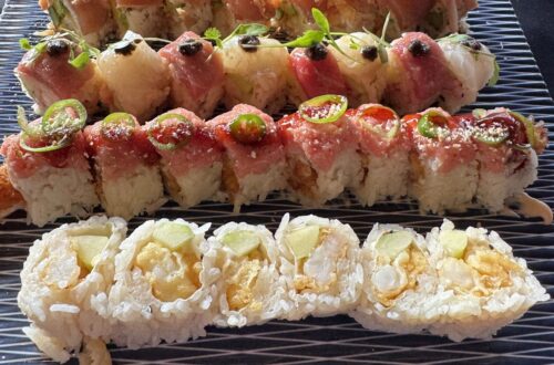 An overhead view of a large platter featuring four varieties of specialty sushi rolls, including the Tokubetsu Roll topped with delicate microgreens and black accents, the White Lotus Roll, a spicy tuna roll with jalapeño slices, and a popcorn shrimp tempura roll, served on a textured blue and white glass plate at Sushi Roku in Santa Monica.