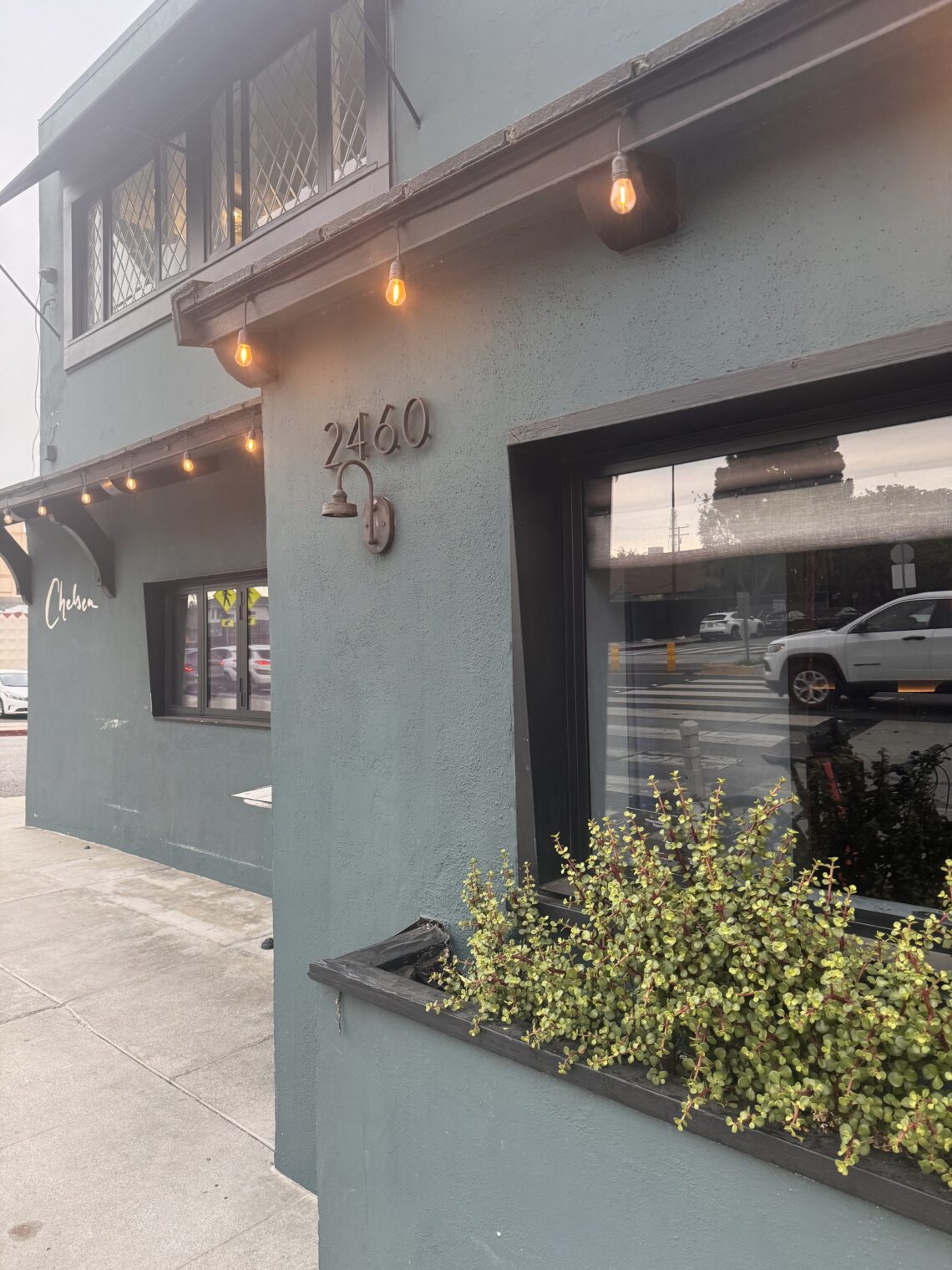 A street-level, angled view of the exterior of Chelsea Restaurant. The two-story building features a matte, dark slate-gray stucco facade. On the main corner wall, the house number "2460" is mounted in dark metal above a matching gooseneck light fixture. Warm, amber bistro string lights hang under the roofline eaves. A large glass window reflects the street outside, while a black planter box beneath it overflows with small-leafed green succulents. Further down the sidewalk, the word "Chelsea" is written in elegant, white cursive script on the building's wall.